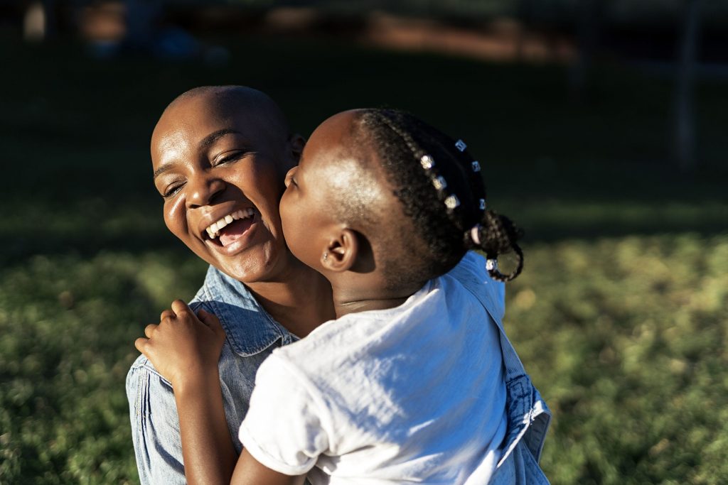 A kid with a fade and braids kisses their mother on the cheek. She is laughing in delight.