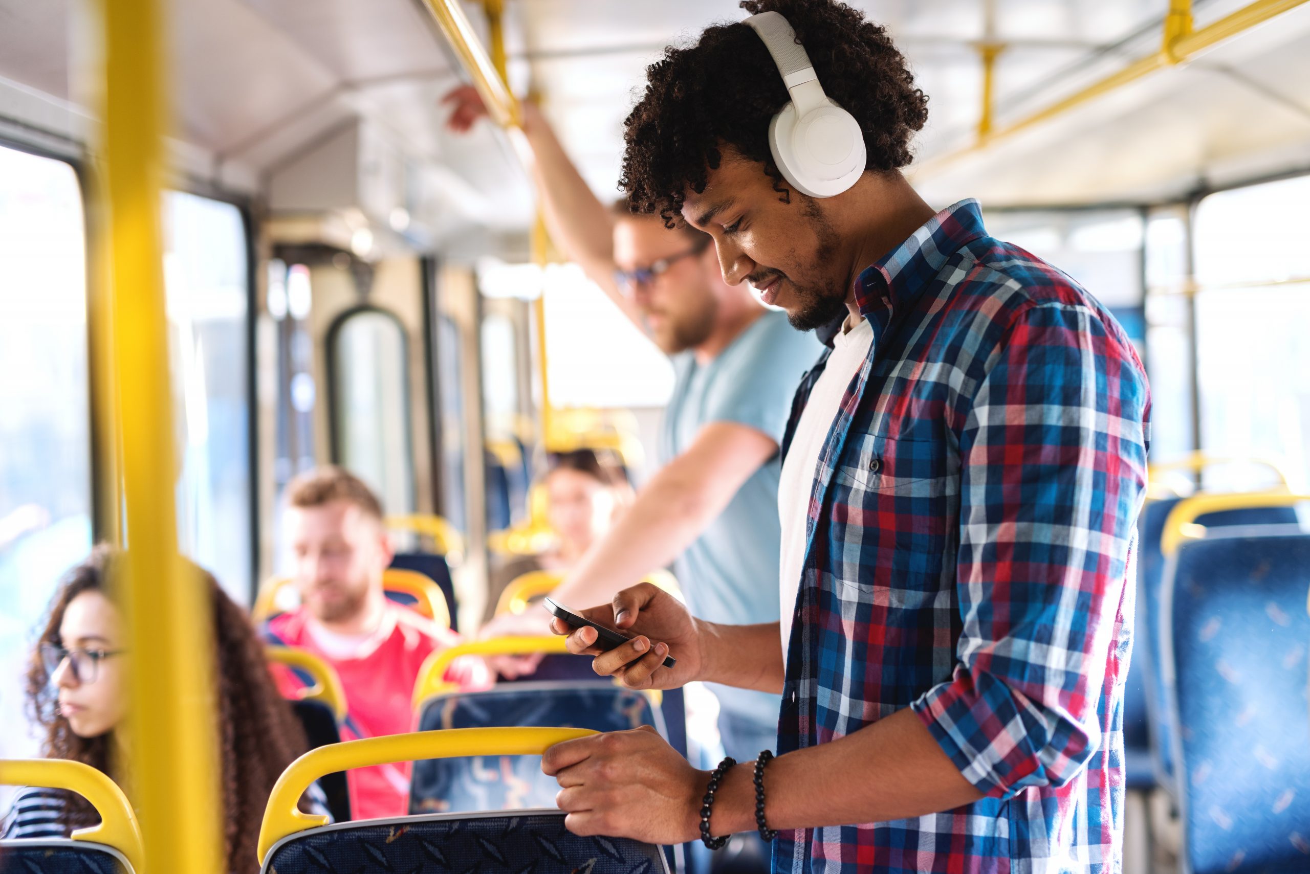 A man wearing a plaid shirt and headphones looks down at his phone while riding the bus.