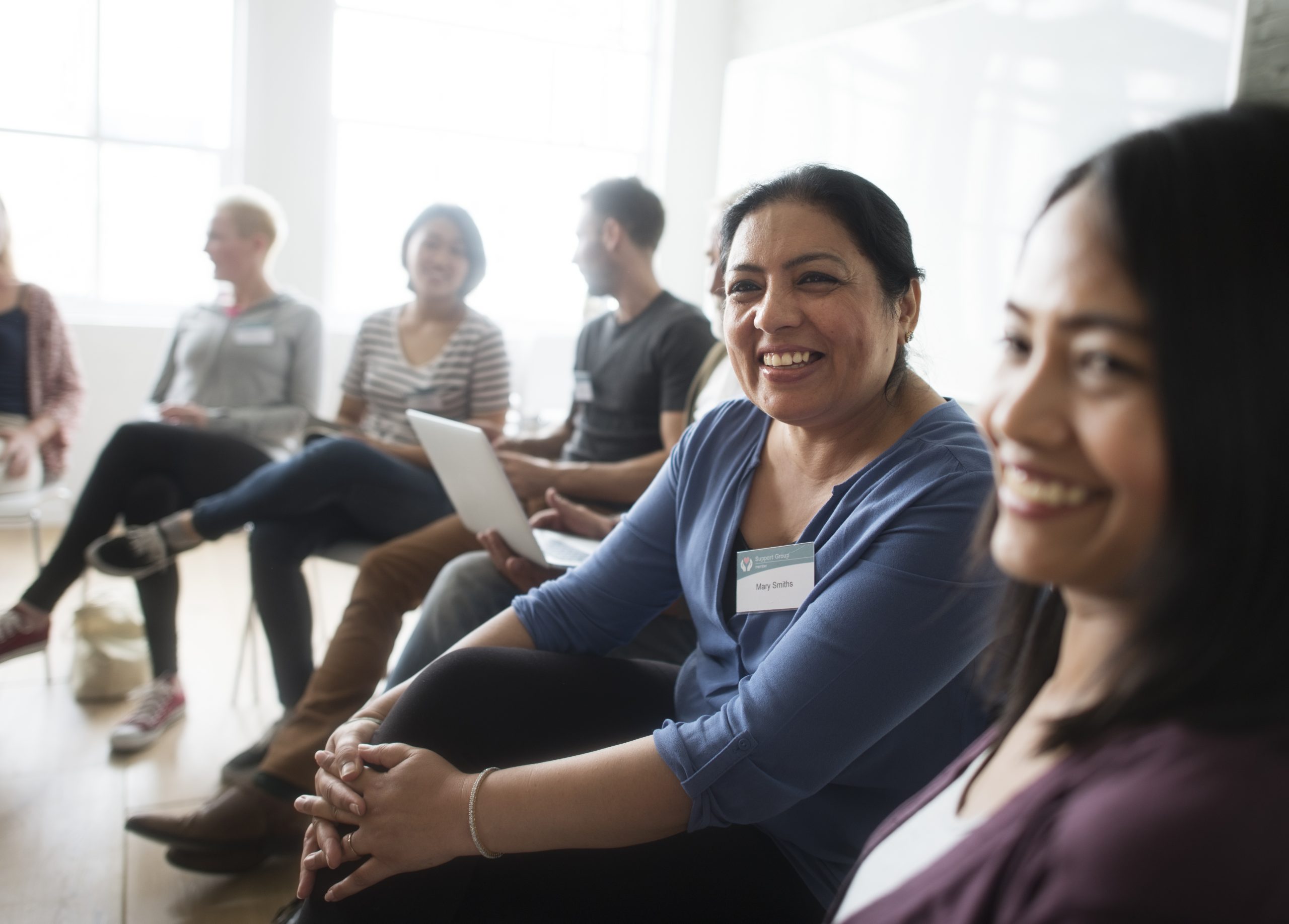 A woman sits in a community meeting, smiling at the camera.