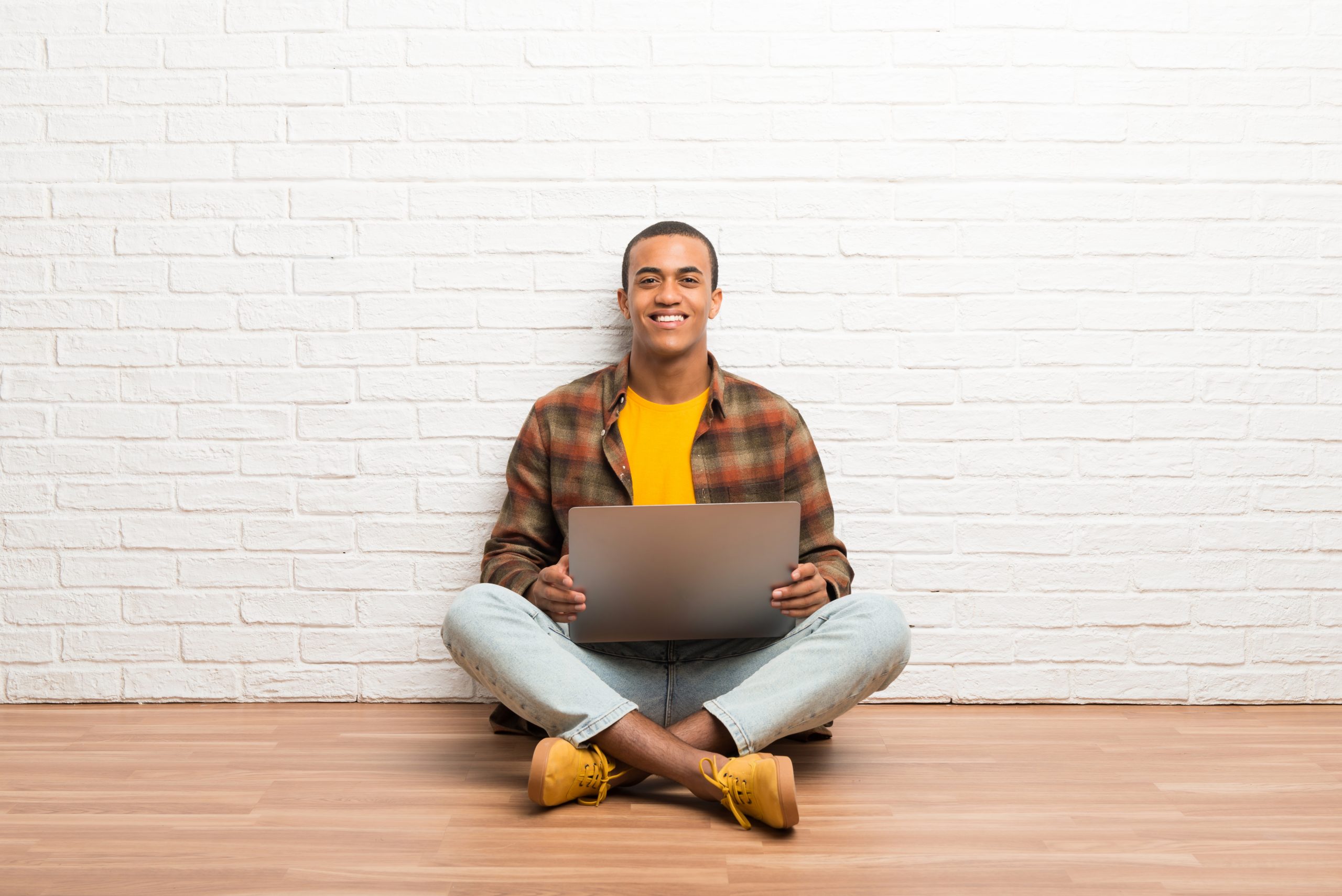 A young black man sits with his legs crossed holding his laptop in front of a white brick wall.