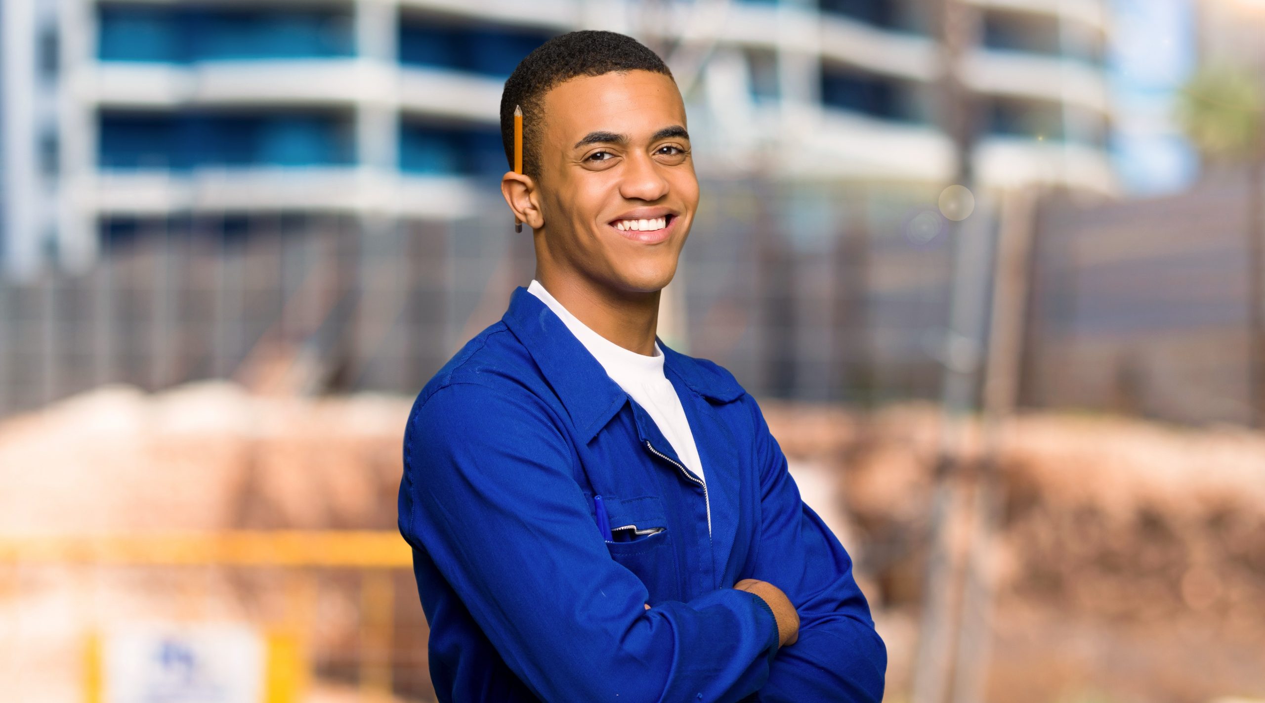The young man stands smiling with his arms crossed wearing a smart blue work shirt.