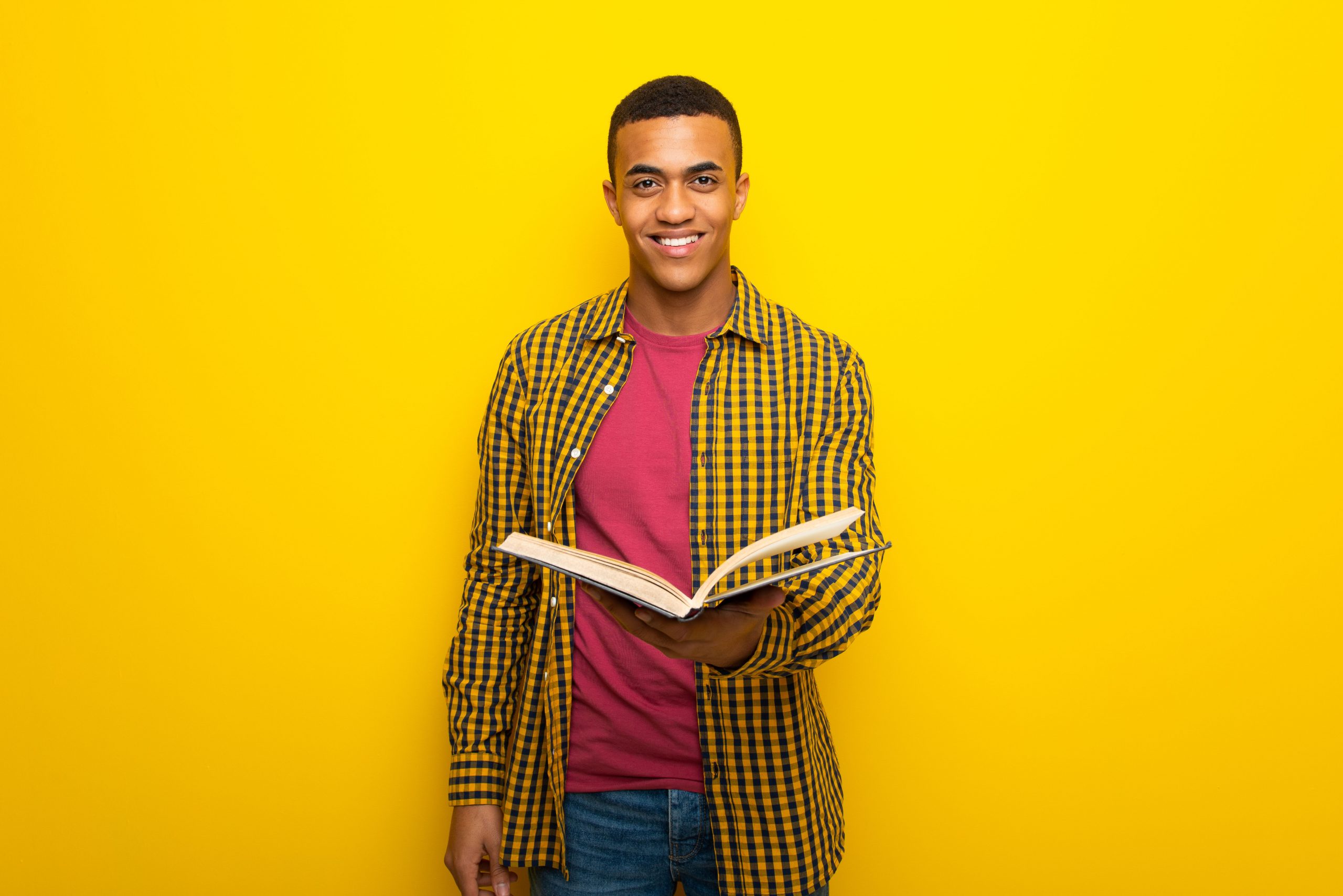 The young man is holding a textbook and standing against a yellow background.