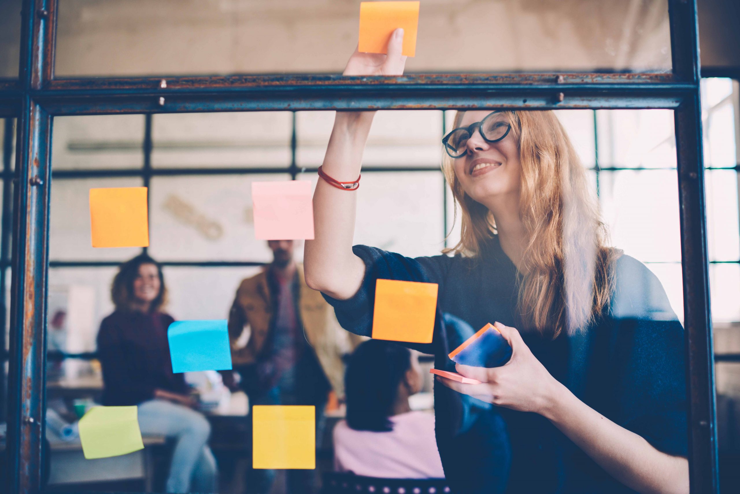 A red haired woman adds an orange Post-It note to a window while her cowrokers brainstorm.