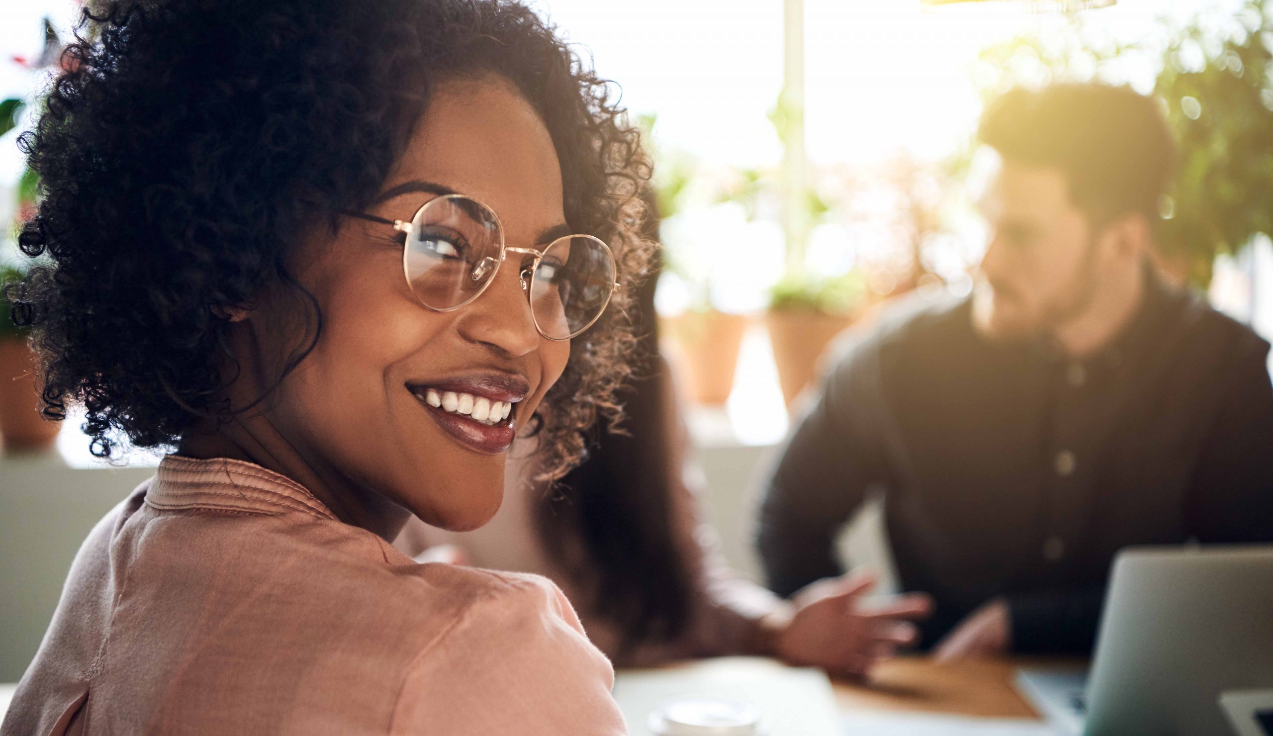 A black woman wearing glasses turns to look at the camera while sitting at a conference table with her peers.