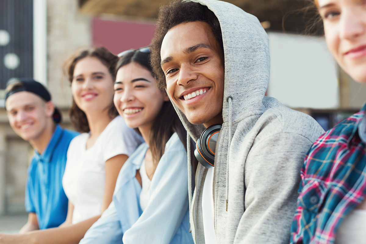 A group of diverse teenagers sit together on a city bench. The image is focused on a Black boy in a grey hoodie.