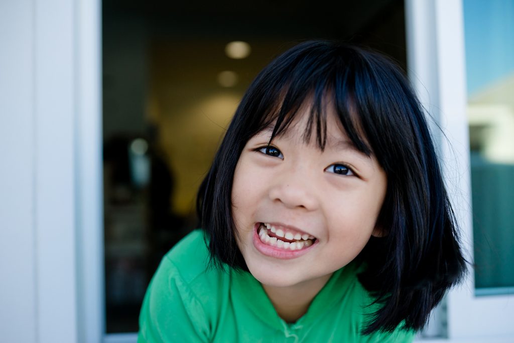 A little girl in a green shirt smiles, leaning out a window on a sunny day.