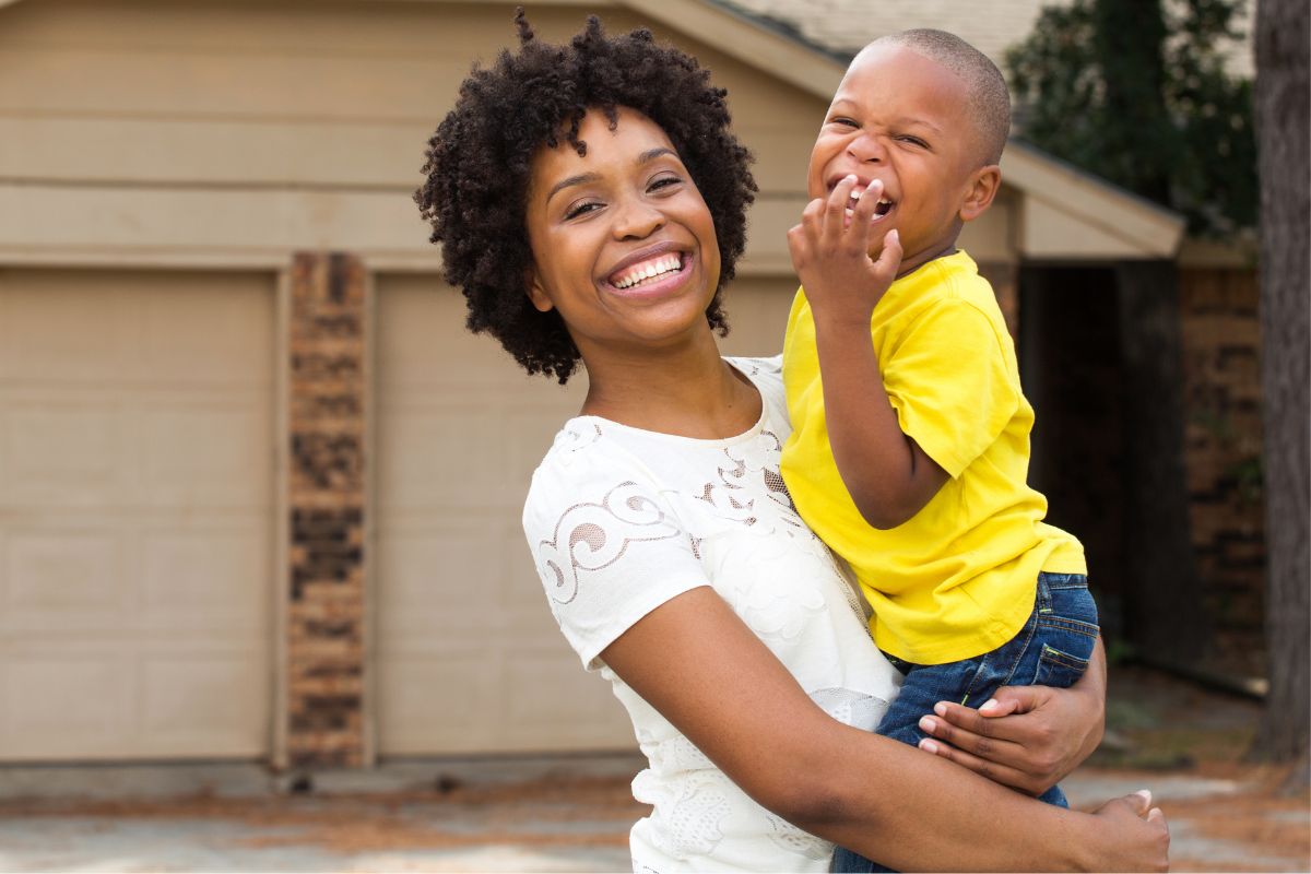 A Black mom and her toddler son smile in front of their new house.