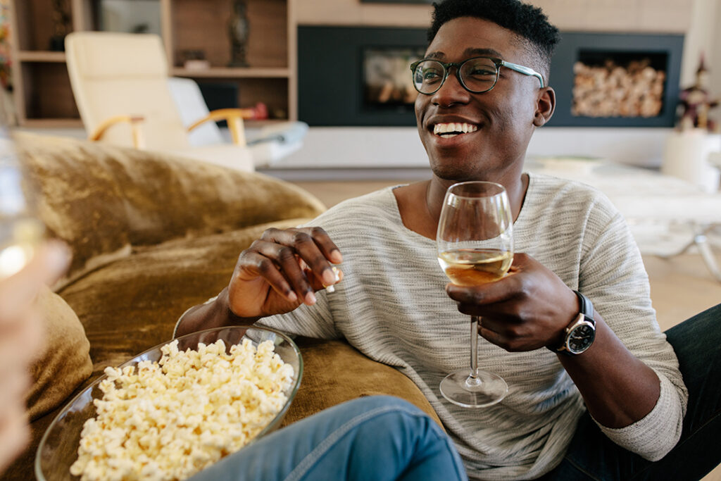 Black man sitting by a sofa with glass of wine and eating popcorn.