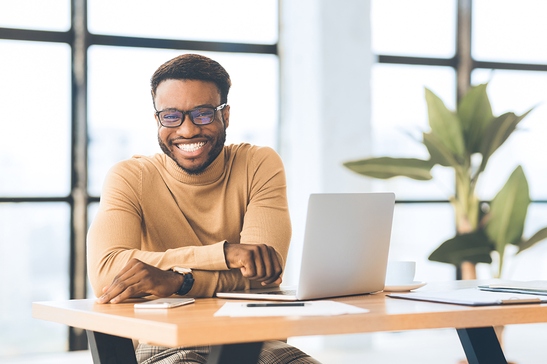Smiling black man looking at camera, sitting at desk with laptop