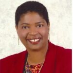 Dr. Jamilah R. Jor'dan Photo Jamilah is a Black woman with short hair and a big smile wearing a red blazer.